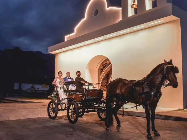 Novios en un carruaje tirado por caballo frente a la capilla iluminada de Hacienda Las Marías, boda destino cerca de Bogotá